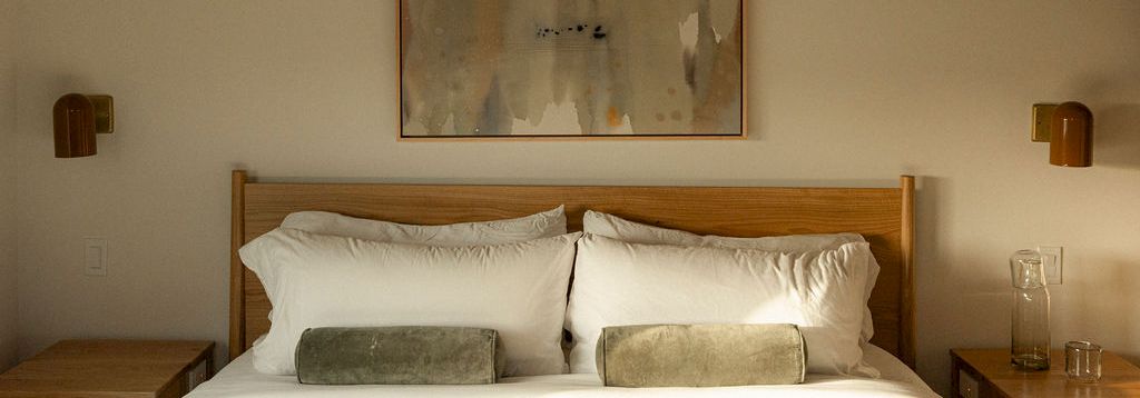 A tidy hotel bedroom with a wooden bed, white linens, two small pillows, wall art above, and simple bedside tables on either side.