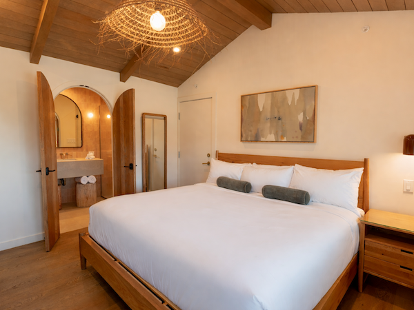 A bright, cozy bedroom with a large bed, wooden furniture, arched doorway to a bathroom, warm lighting, and a woven pendant-style ceiling light.