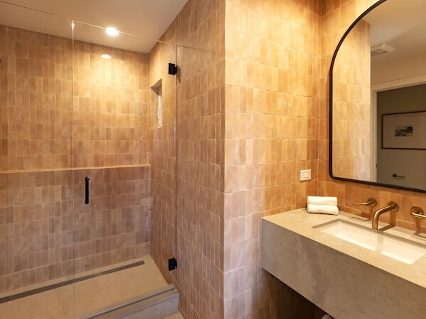 A modern bathroom with a walk-in shower, tan tiled walls, a double sink vanity, and a large arched mirror; bright and clean.