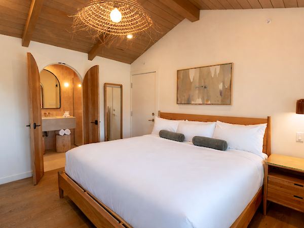 A bright, cozy hotel bedroom with a large wooden bed, white linens, two green bolster pillows, wooden nightstands, and an open doorway leading to a tiled ensuite.