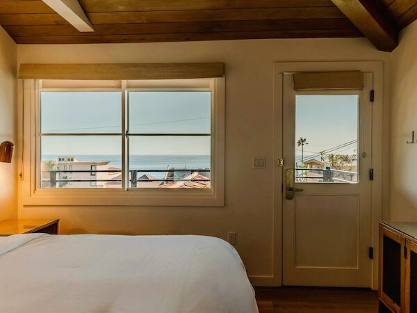 A bright hotel room with a bed, nightstand, window view of rooftops and the sea, and a door leading to a balcony or hallway.