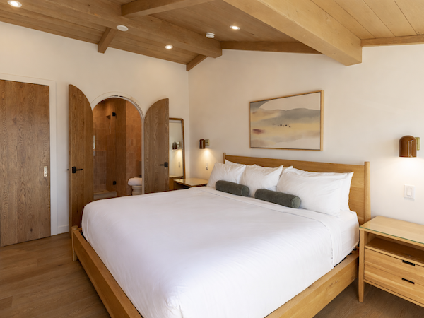 A spacious bedroom with a wooden bed, white bedding, and matching nightstands; exposed wooden beams, arched doors, and soft wall art.