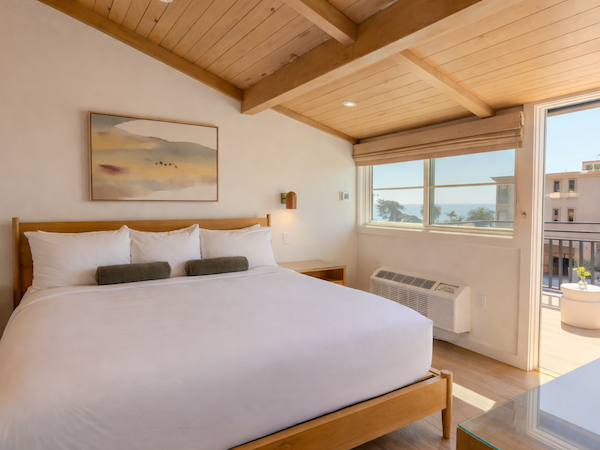 A bright hotel room with a large white bed, wooden frame, soft pillows, a painting above, and a sunny balcony with a view of the city.