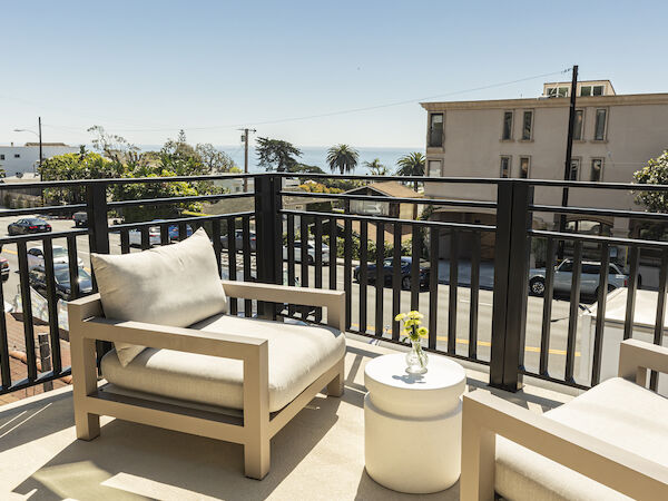 A sunny balcony with beige outdoor seating and a small round table, overlooking a street and distant buildings, under a clear blue sky.