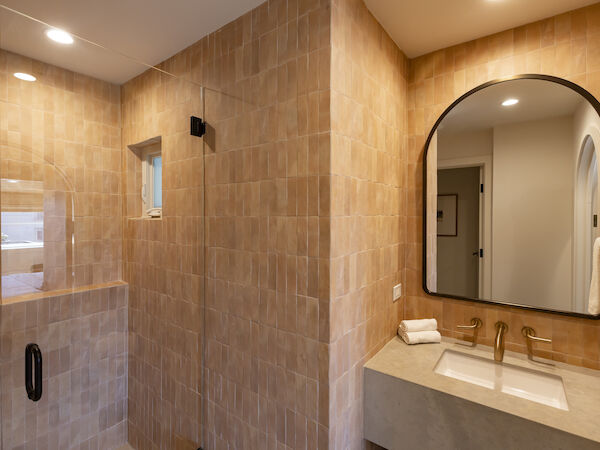 A modern bathroom with tan tiled walls, a glass shower, and a rounded mirror above a double sink; warm lighting completes the space.