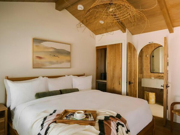 A cozy hotel room with a large bed, white linens, wooden accents, and a small breakfast tray on the bed near a doorway to a bathroom.