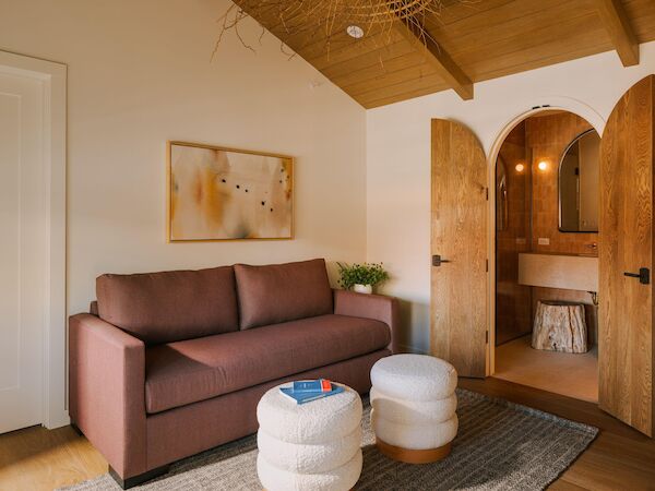 A cozy living space with a brown sofa, two white tufted ottomans, a woven rug, and a wooden arch doorway leading to a bright area.
