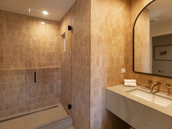 A modern bathroom with a glass-enclosed shower on the left, beige tiled walls, a double sink with a large rounded mirror on the right, and stacked towels by the sink.