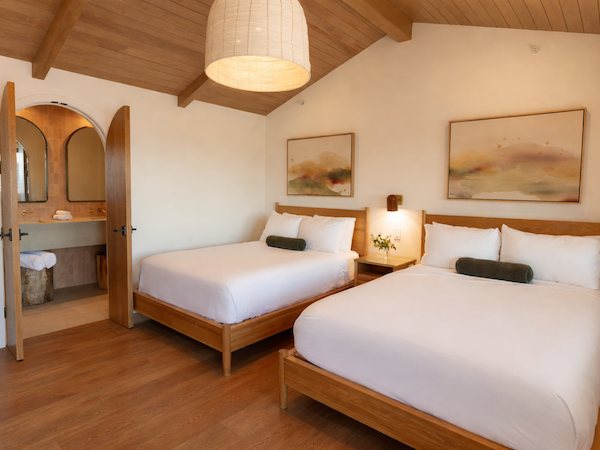 A cozy hotel room with two single beds and a shared wooden headboard, warm lighting, a ceiling lamp, and an open doorway to a small bathroom.