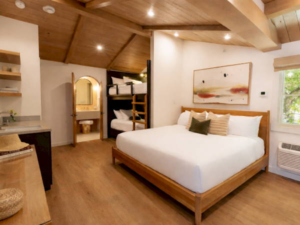 A cozy wooden bedroom with a large bed, bunk-style closet, warm lighting, and a small desk; rustic ceiling beams and a window overlook greenery.