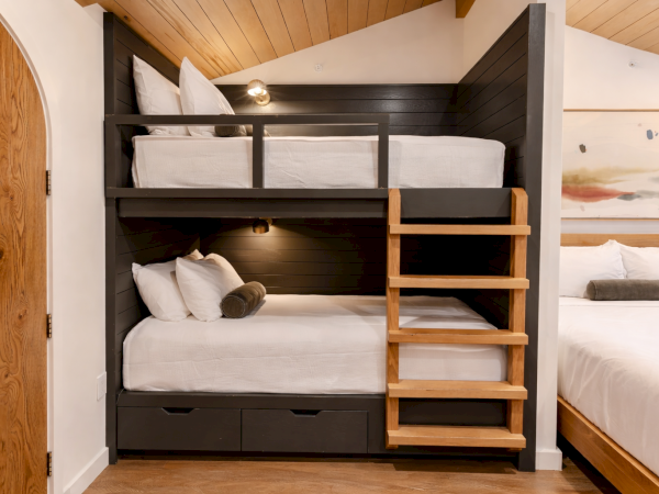 A cozy bedroom with a dark wooden bunk bed and ladder, white bedding, soft pillows, a wooden ceiling, and a separate full bed nearby, all in a compact space.