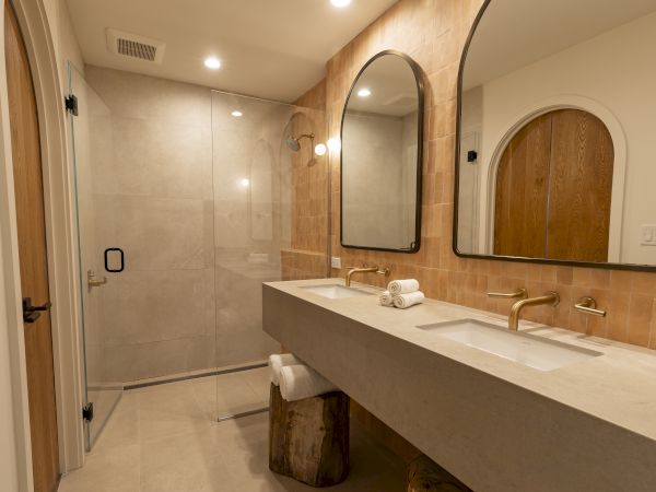 A modern bathroom with a double vanity, two rectangular mirrors, warm beige tiles, a glass walk-in shower, and wood accents, bright lighting.