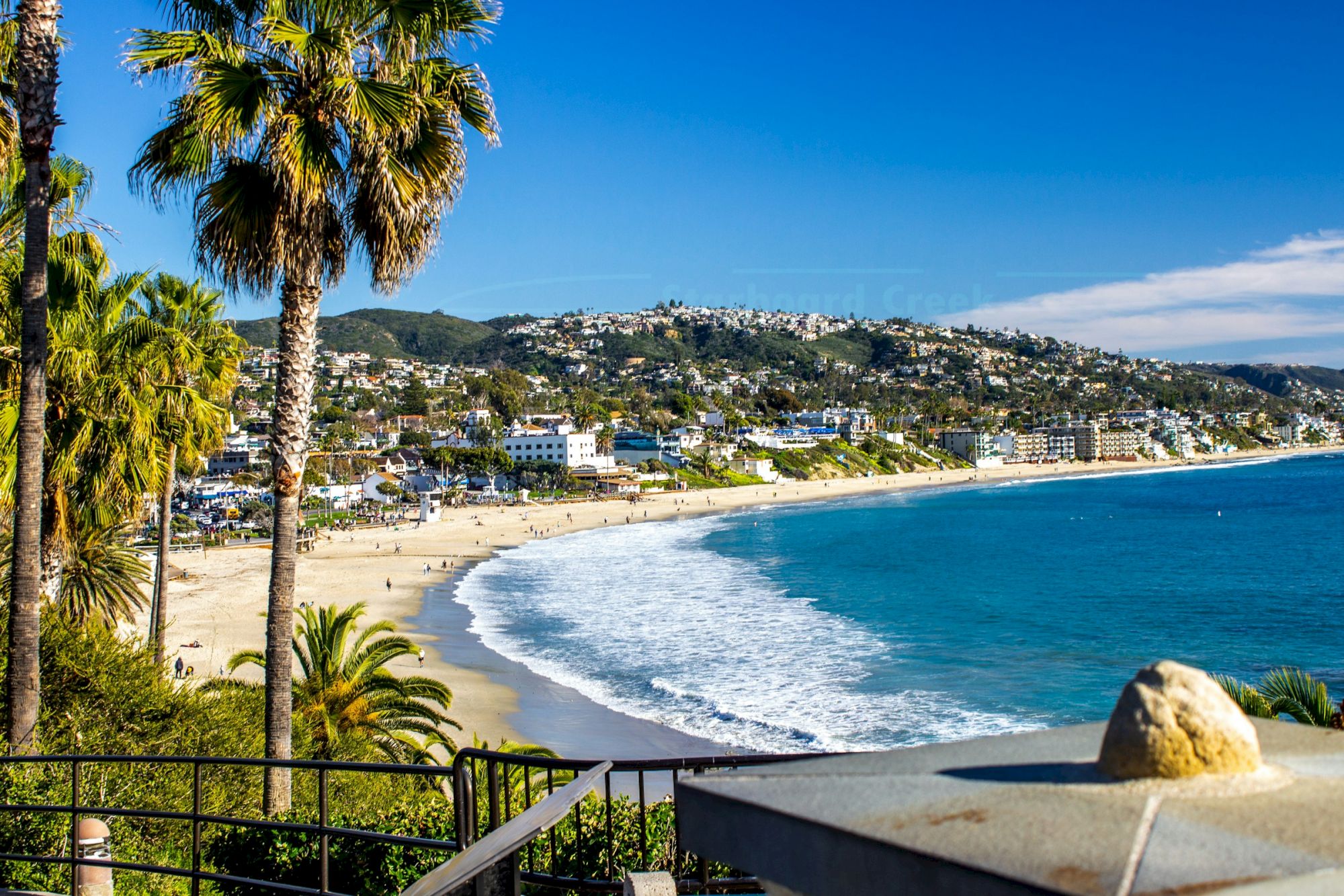 A sunny coastal scene with a curved beach, palm trees, blue sea, and a hillside town in the distance, bright and inviting.