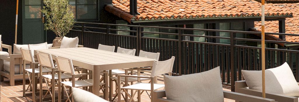 A sunny rooftop patio with white tables, chairs, and umbrellas, bordered by a black railing and terracotta-tiled roofs, palm trees in the background.