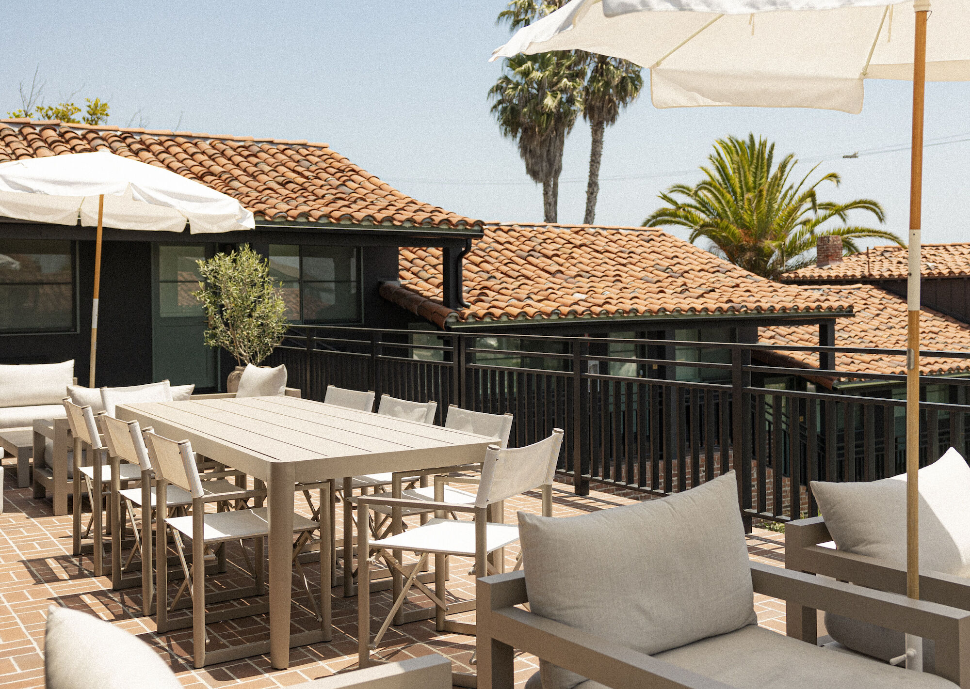A sunny rooftop patio with white furniture, tables and chairs, beige umbrellas, potted plants, and terracotta-tiled roofs in the background.