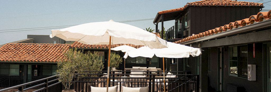 A sunny rooftop lounge with white cushioned seating, umbrellas, and a railing overlooking neighboring buildings.