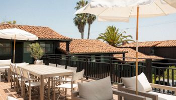 Outdoor resort patio with white cushioned seating, tables, umbrellas, and palm trees in the background, sunny and relaxing ambiance.