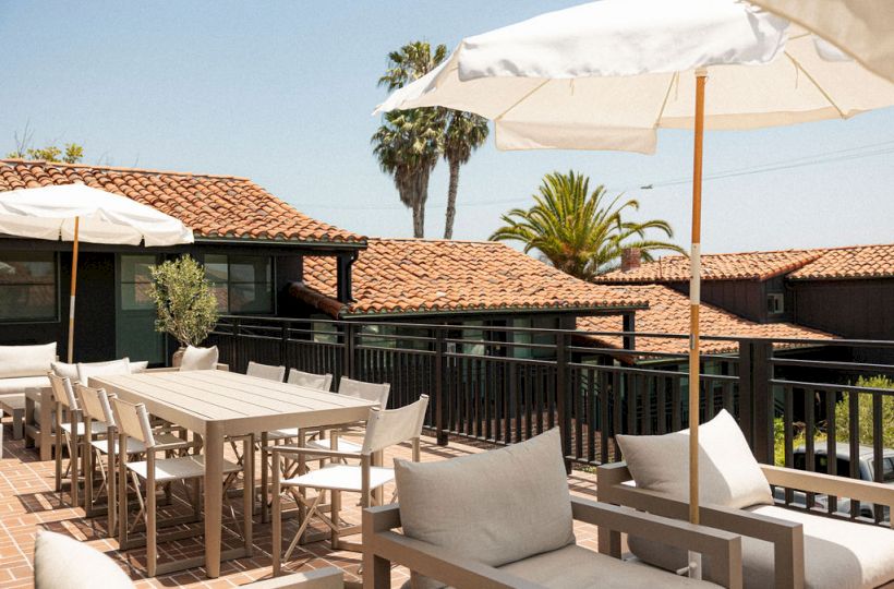 Outdoor resort patio with white cushioned seating, tables, umbrellas, and palm trees in the background, sunny and relaxing ambiance.