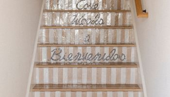 Staircase with striped carpet runners, a beige rug at the bottom, white walls, a hat on the left, and &ldquo;Bienvenidos&rdquo; written across the steps.