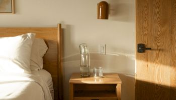 A simple hotel room: a wooden bed, white sheets, a nightstand with a glass water bottle and a shot glass, a wall-mounted lamp, and a wooden door.