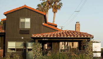 A two-story building with a terra-cotta roof, beige and dark siding, palm trees behind, a small garden, and a sign near the entrance.