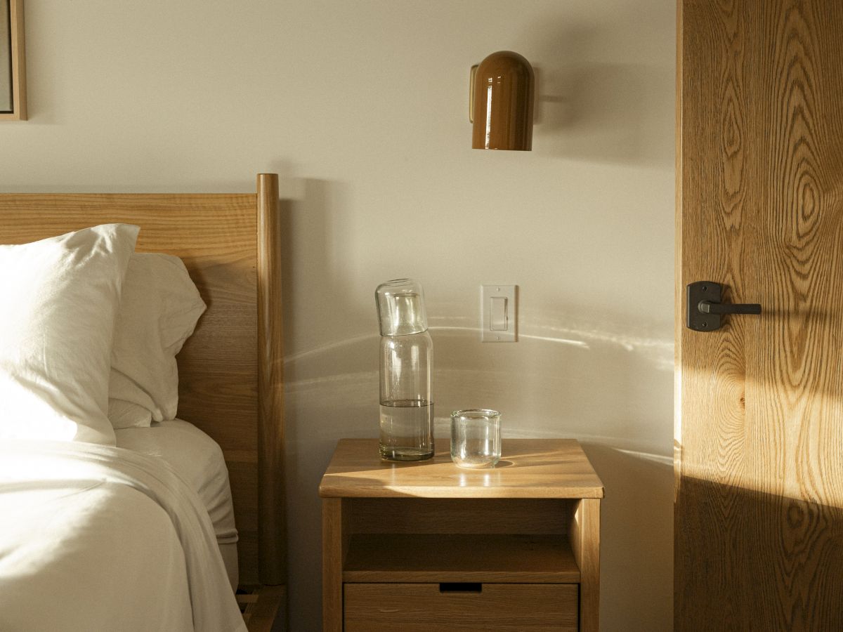 A tidy hotel bedside scene with a wooden headboard, white bed linens, a small wooden nightstand, a glass of water, a door, and a wall-mounted lamp.
