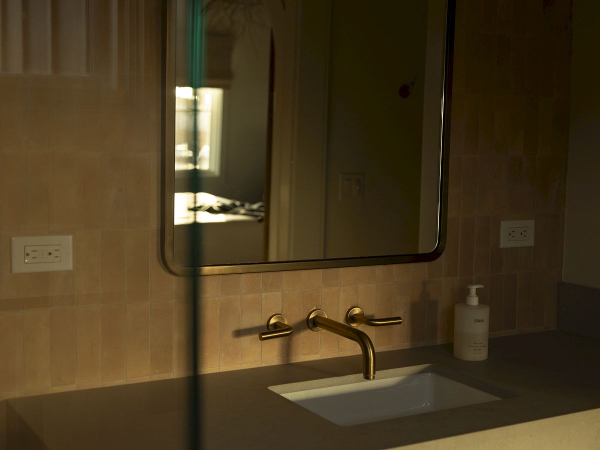 A bathroom sink with a modern faucet, mirror, and beige tiled walls; soft lighting creates a calm, dim atmosphere.