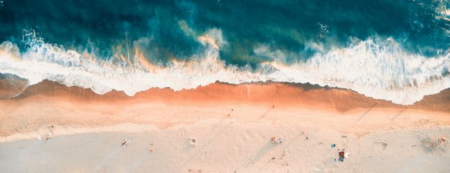 Aerial view of a tropical beach: turquoise waves meet pale sand, foamy white crests kiss the shore, sunlight warms the tranquil shoreline.