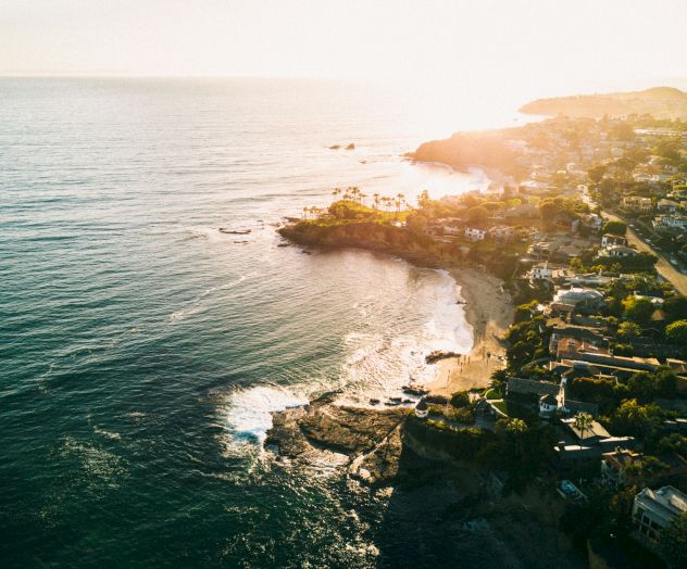 A coastal town on rocky cliffs with sunlight kissing the shoreline, waves crashing, and homes cascading along the seaside, serene and sunny.