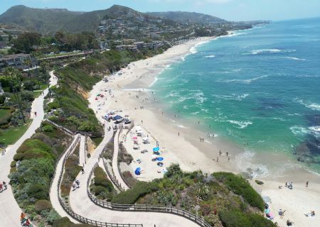 Aerial view of a sunny beach with turquoise water, winding boardwalks, cottages on cliffs, and people sunbathing and strolling along the shore.