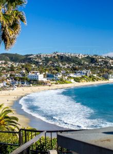 A sunny coastal scene with a crescent beach, blue ocean, palm trees, and a hillside town in the distance, bright and vibrant.