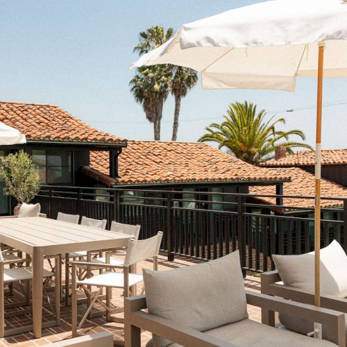 A sunlit outdoor dining area with wooden tables, white chairs, large umbrellas, and a calm courtyard setting.
