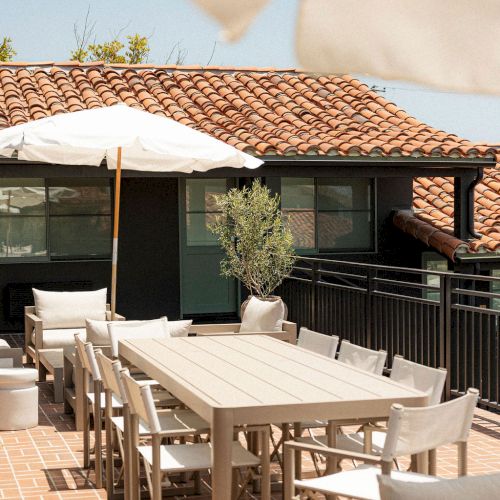 An outdoor dining area with a long wooden table, chairs, and a shaded patio under a tiled roof. End of sentence.