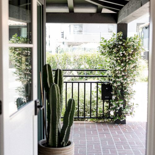 A small, shaded porch with a tiled floor, potted plants, a wooden door, and a garden view beyond the railing.