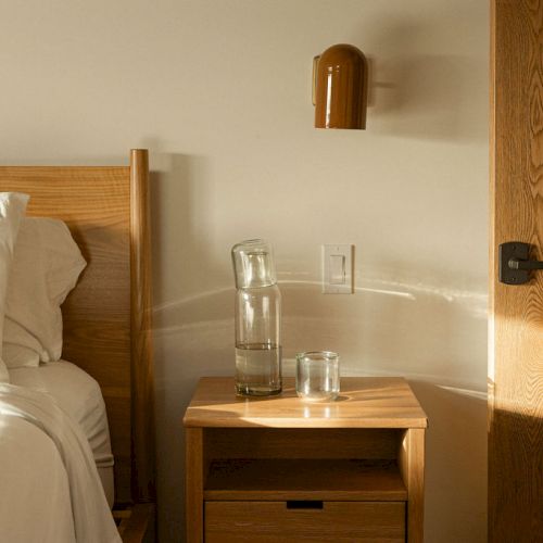 A cozy bedroom with a single bed, wooden nightstand, a small shelf, warm lighting, and a rustic wall with a small window.