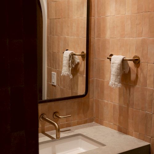 A small bathroom vanity with a sink, mirror, and tissue holder; warm brown tiles and soft lighting, a minimalist setup.