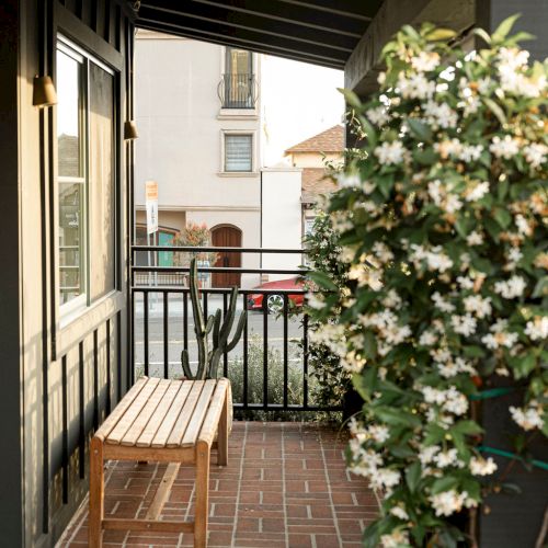 A cozy brick balcony with a small wooden bench, potted flowers, and a narrow path leading to a neighboring building.