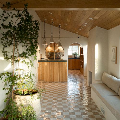 A sunny hallway with a tiled floor, wooden beams, potted plants, and a bright kitchen area at the end, light streaming through windows.