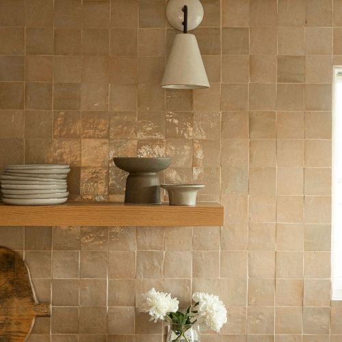 A cozy kitchen shelf scene with a brick wall, hanging metal light fixture, wooden shelves, jars and bowls, and a small bouquet on the counter.