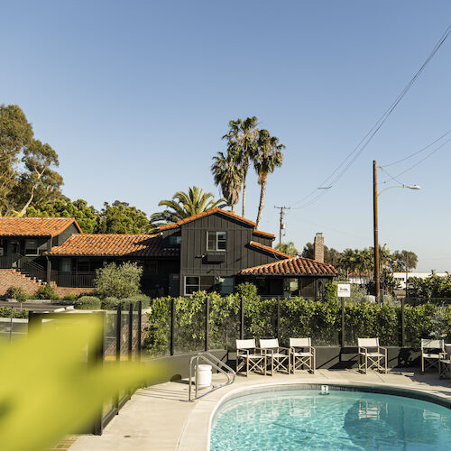 Outdoor resort scene with a swimming pool, patio chairs, tiled roofs, palm trees, and a bright blue sky with overhead wires.