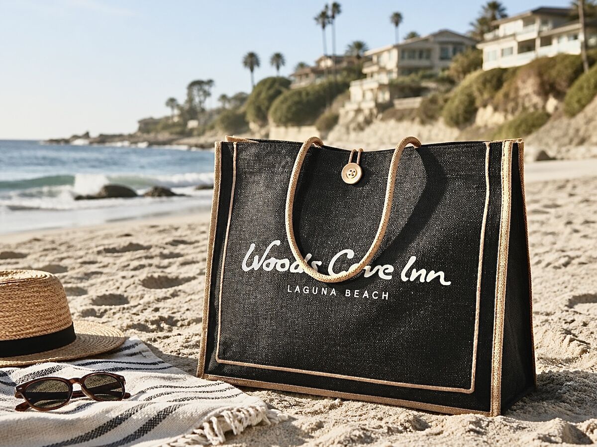 A beach scene with a dark tote bag reading &ldquo;Woodstone Inn Laguna Beach&rdquo; on the sand, along with sunglasses and a straw hat nearby.