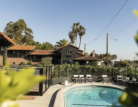 A sunny pool courtyard with lounge chairs, tiled-roof buildings, palm trees, and power lines in the background.