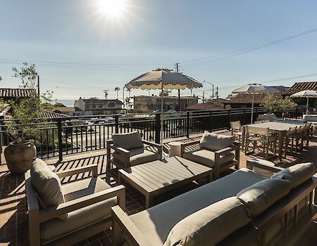 Sunny rooftop terrace with cushioned loungers, outdoor table, and striped umbrellas, overlooking houses and the coast.