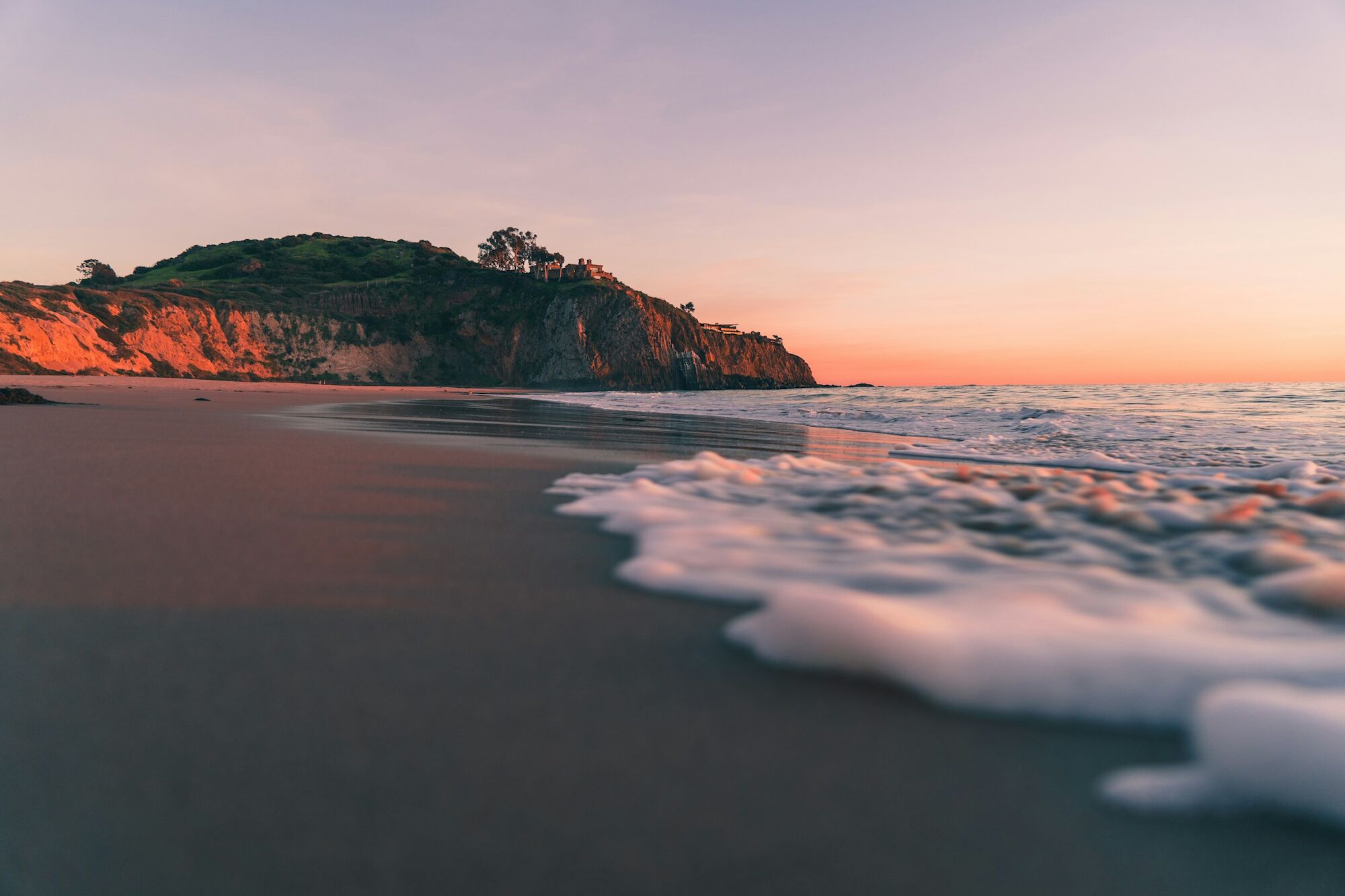 A serene sunset over a sandy beach, with waves rolling in and a cliff-top house glowing on the horizon.