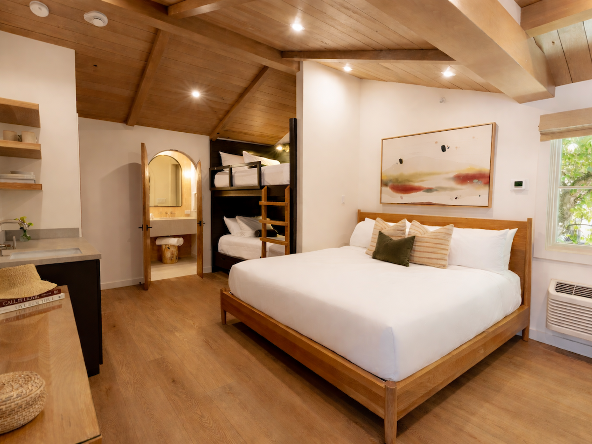A bright modern bedroom with a wooden ceiling, large bed with pillows, wall art, open doorway to a bathroom, and shelving.