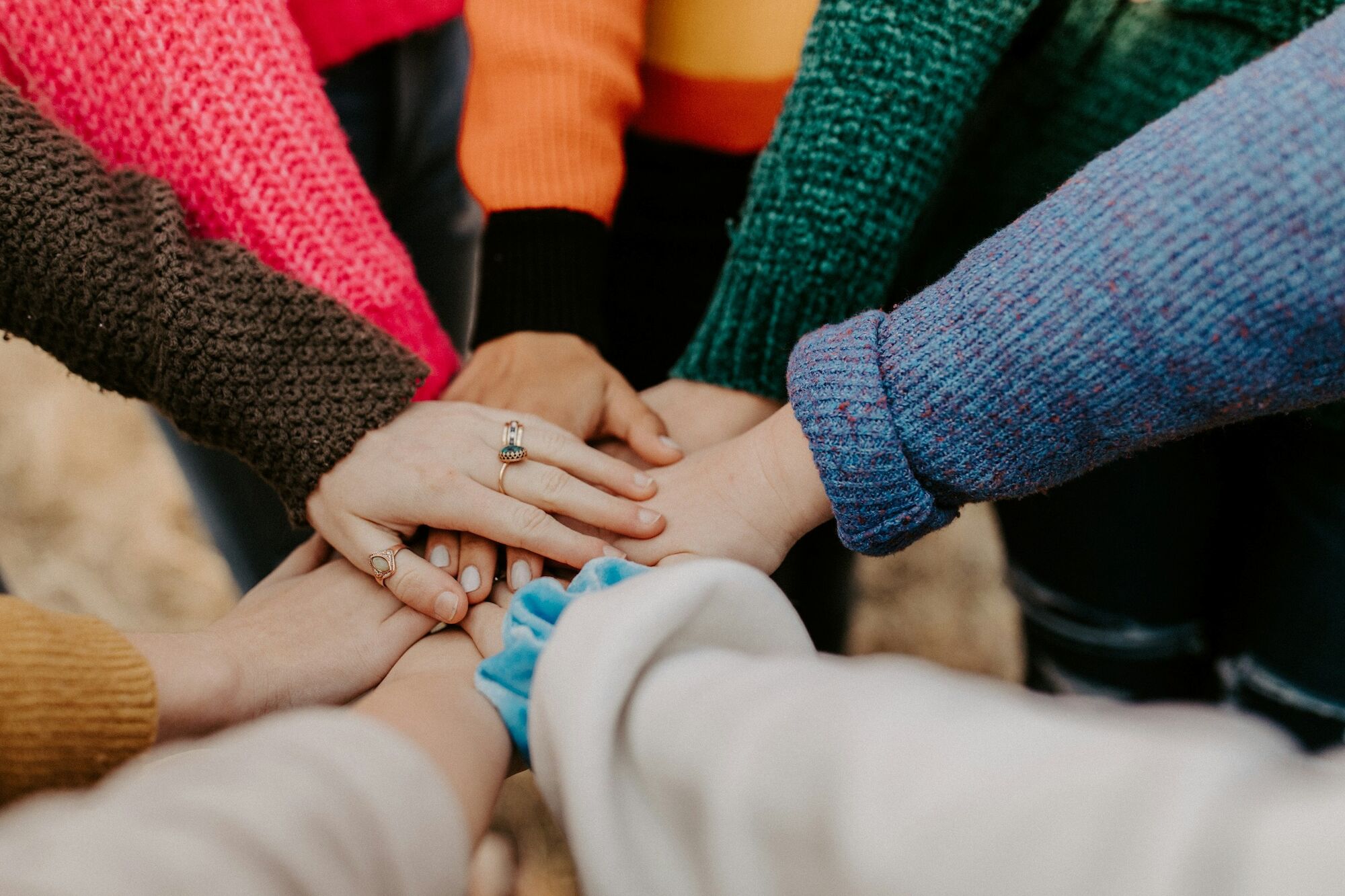 Several people&rsquo;s hands with rings gently cradle a baby as knit sleeves form a loving circle around it.