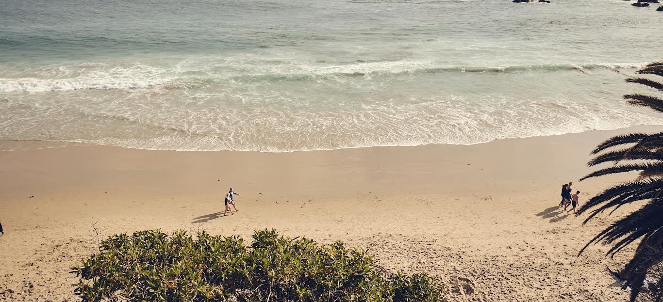 Sunny beach with gentle waves, palm trees, and people walking and picnicking along the shoreline.