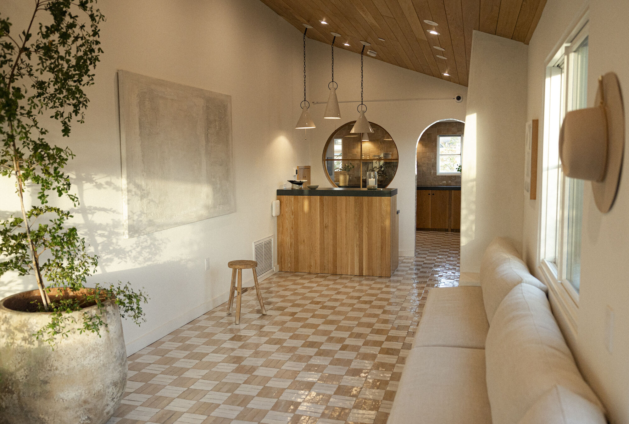 Modern indoor kitchen area with wood cabinetry, hanging pendant lights, a round mirror, tiled floor, and patio greenery outside.