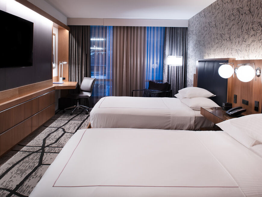 A modern hotel room with two beds, wood paneling, TV on the wall, desk, and dark curtains by large windows.