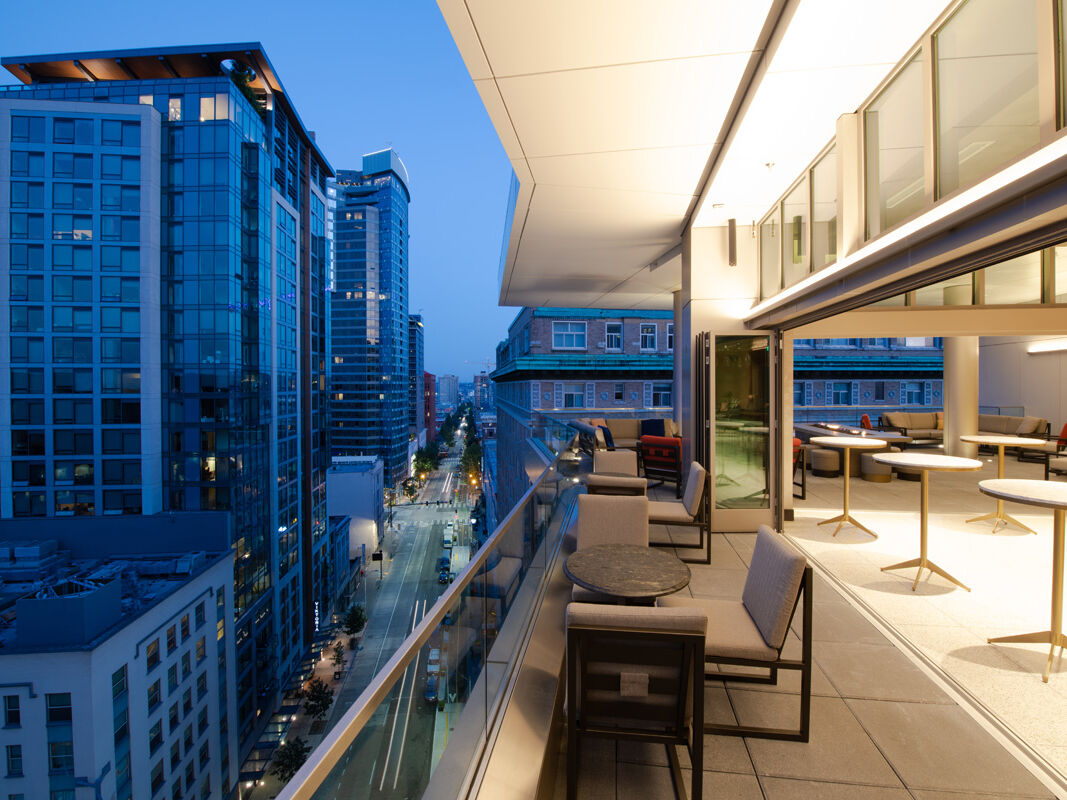 Modern high-rise skyline view from a rooftop terrace with lounge chairs overlooking a city street at dusk.
